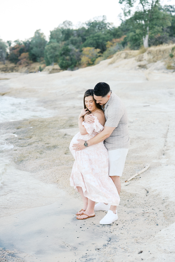 Pregnant woman in a pink maternity gown standing at the water's edge at Folsom Lake